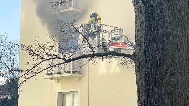 © Feuerwehr Dresden Frau steht auf Balkon von qualmender Wohnung. Drehleiter mit einem Feuerwehrmann fährt in Stellung