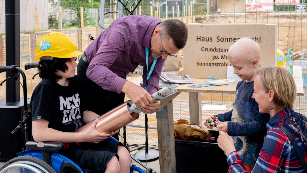 auf dem Bild füllen der fünfjährige Fabian (im Bild mit seiner Mama) und  der 12jährige Adam gemeinsam mit Falk Noack vom Sonnenstrahl e.V. die Zeitkapsel, die in den Grundstein eingemörtelt wird © Kirsten Lassig